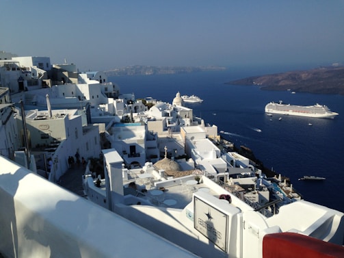 A picturesque coastal town with white buildings perched on a hillside overlooking a deep blue sea. The architecture is traditional, with cubic houses and narrow paths. Cruise ships are visible in the water, suggesting a popular tourist destination.