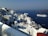 A picturesque coastal town with white buildings perched on a hillside overlooking a deep blue sea. The architecture is traditional, with cubic houses and narrow paths. Cruise ships are visible in the water, suggesting a popular tourist destination.
