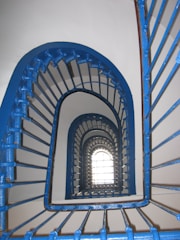 A spiral staircase with blue railings creates an optical illusion effect as it ascends. The pattern of the metal bars adds a rhythmic, geometric design, leading the eye upwards towards a bright window at the top.