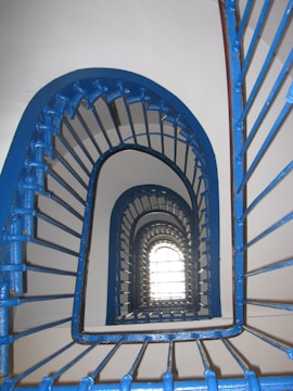 A spiral staircase with blue railings creates an optical illusion effect as it ascends. The pattern of the metal bars adds a rhythmic, geometric design, leading the eye upwards towards a bright window at the top.