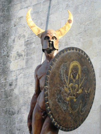 A statue of a warrior with a golden helmet adorned with large curved horns. The figure holds a large, ornate shield decorated with a mythical creature. The background consists of a stone wall with visible block patterns, suggesting an outdoor setting.