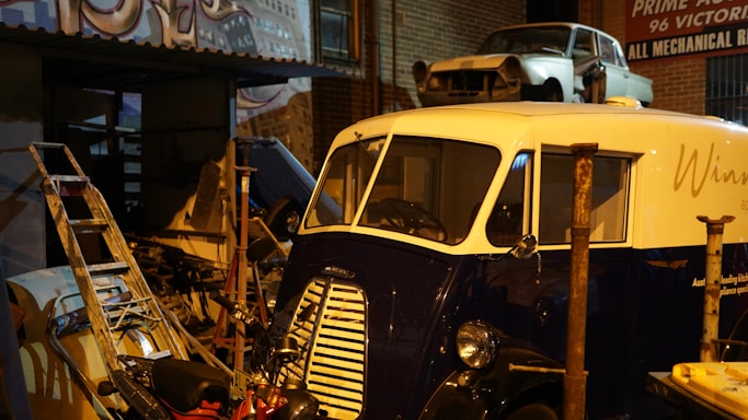 A vintage vehicle with a blue and white paint job is parked in what appears to be a garage or workshop. Around the vehicle, there are various tools and items including a ladder and a motorcycle. On a platform above, there is another disassembled car. The setting is dimly lit, contributing to an old-fashioned and somewhat cluttered ambiance.