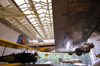 Aviation exhibit featuring several airplanes displayed indoors. A large metal wing dominates the right side of the frame, reflecting the light filtering through the building’s glass ceiling. Vintage planes with 'Fly East' and 'Eastern Air Transport' written on them are visible, showing the history of air travel.