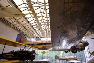 Aviation exhibit featuring several airplanes displayed indoors. A large metal wing dominates the right side of the frame, reflecting the light filtering through the building’s glass ceiling. Vintage planes with 'Fly East' and 'Eastern Air Transport' written on them are visible, showing the history of air travel.