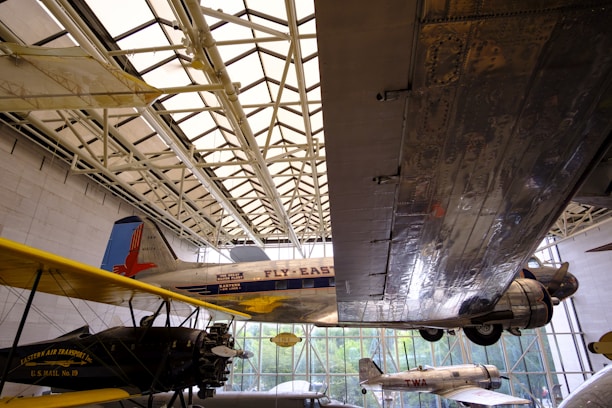 Aviation exhibit featuring several airplanes displayed indoors. A large metal wing dominates the right side of the frame, reflecting the light filtering through the building’s glass ceiling. Vintage planes with 'Fly East' and 'Eastern Air Transport' written on them are visible, showing the history of air travel.
