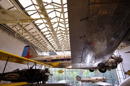 Aviation exhibit featuring several airplanes displayed indoors. A large metal wing dominates the right side of the frame, reflecting the light filtering through the building’s glass ceiling. Vintage planes with 'Fly East' and 'Eastern Air Transport' written on them are visible, showing the history of air travel.