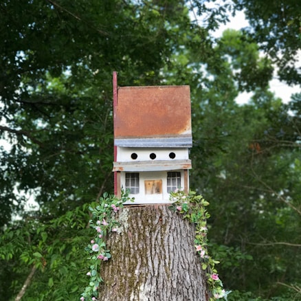 A birdhouse with a weathered, rust-colored roof sits atop a tree stump. The birdhouse has multiple circular openings and small windows. It is decorated with a garland of pink and white flowers and surrounded by lush green foliage in the background.