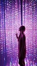 A woman stands in front of a dazzling array of LED lights that create a vibrant and immersive atmosphere. The lights form a curtain of pink, purple, and blue hues, with some bulbs varying in brightness to add depth and interest.