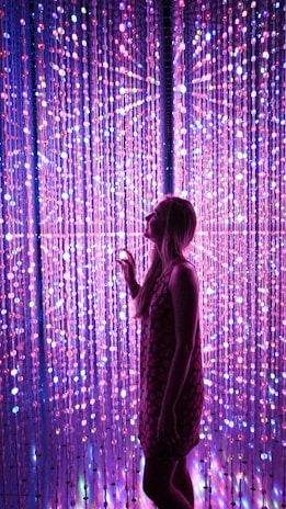A woman stands in front of a dazzling array of LED lights that create a vibrant and immersive atmosphere. The lights form a curtain of pink, purple, and blue hues, with some bulbs varying in brightness to add depth and interest.