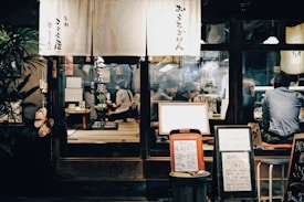 A cozy interior of a traditional Japanese restaurant with people sitting on tatami mats around low tables, enjoying their meals. The entrance is decorated with a curtain displaying Japanese characters, and several menu boards are placed outside, featuring handwritten text. Soft lighting creates an inviting atmosphere.