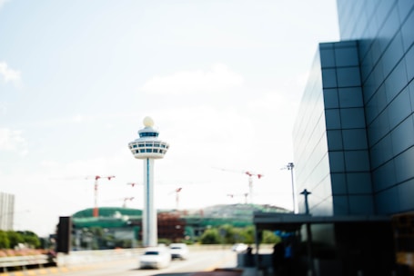A modern control tower stands prominently, contrasting against a pale blue sky. Red cranes and under-construction buildings are visible in the distance, suggesting an active development site. A sleek, glass-clad building on the right adds to the urban atmosphere. The scene is accented by a blurred road below with vehicles in motion.