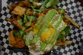 A savory dish featuring a mix of crispy fries topped with a sunny side up egg. Green sauce is drizzled over the dish, which includes spinach leaves and pieces of golden-brown fried chicken.