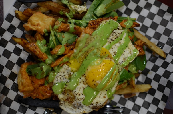 A savory dish featuring a mix of crispy fries topped with a sunny side up egg. Green sauce is drizzled over the dish, which includes spinach leaves and pieces of golden-brown fried chicken.
