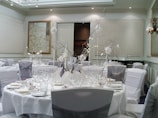 A well-decorated banquet hall with elegantly set round tables. The tables are covered with white tablecloths and adorned with sophisticated centerpieces featuring branches and white accents. Elegant place settings including plates, glasses, and cutlery are meticulously arranged. There are cushioned chairs covered with white and silver chair covers surrounding each table. The walls have a refined design with neutral tones and framed artwork, and the ceiling sports recessed lighting and decorative molding.