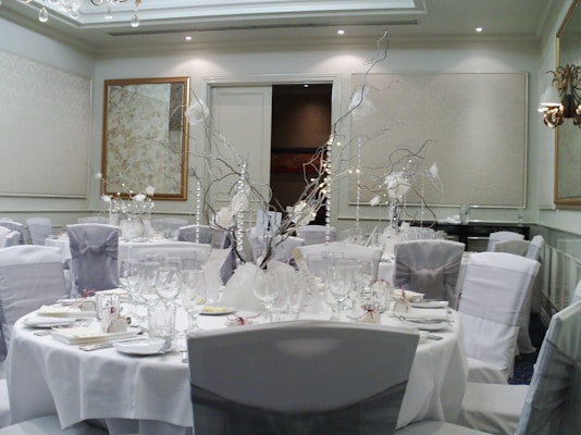 A well-decorated banquet hall with elegantly set round tables. The tables are covered with white tablecloths and adorned with sophisticated centerpieces featuring branches and white accents. Elegant place settings including plates, glasses, and cutlery are meticulously arranged. There are cushioned chairs covered with white and silver chair covers surrounding each table. The walls have a refined design with neutral tones and framed artwork, and the ceiling sports recessed lighting and decorative molding.