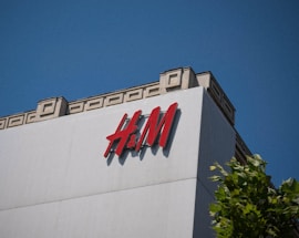A large building featuring the H&M logo prominently on the facade, with a clear blue sky in the background. The structure is modern with geometric designs on the upper edge, and a tree with green leaves is partially visible at the bottom right.