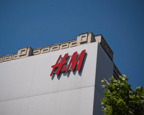 A large building featuring the H&M logo prominently on the facade, with a clear blue sky in the background. The structure is modern with geometric designs on the upper edge, and a tree with green leaves is partially visible at the bottom right.