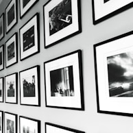 A wall display of framed black and white photographs arranged in a grid pattern. The frames have a sleek, minimalistic design with white mats, and the photos show various scenes, possibly landscapes or candid moments. The arrangement provides a sense of order and adds to the aesthetic appeal of the space.