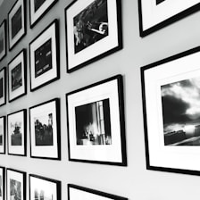 A wall display of framed black and white photographs arranged in a grid pattern. The frames have a sleek, minimalistic design with white mats, and the photos show various scenes, possibly landscapes or candid moments. The arrangement provides a sense of order and adds to the aesthetic appeal of the space.