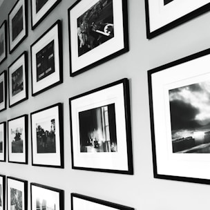 A wall display of framed black and white photographs arranged in a grid pattern. The frames have a sleek, minimalistic design with white mats, and the photos show various scenes, possibly landscapes or candid moments. The arrangement provides a sense of order and adds to the aesthetic appeal of the space.