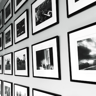 A wall display of framed black and white photographs arranged in a grid pattern. The frames have a sleek, minimalistic design with white mats, and the photos show various scenes, possibly landscapes or candid moments. The arrangement provides a sense of order and adds to the aesthetic appeal of the space.