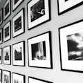 A wall display of framed black and white photographs arranged in a grid pattern. The frames have a sleek, minimalistic design with white mats, and the photos show various scenes, possibly landscapes or candid moments. The arrangement provides a sense of order and adds to the aesthetic appeal of the space.