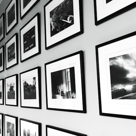 A wall display of framed black and white photographs arranged in a grid pattern. The frames have a sleek, minimalistic design with white mats, and the photos show various scenes, possibly landscapes or candid moments. The arrangement provides a sense of order and adds to the aesthetic appeal of the space.