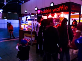 A group of people gathers around a brightly lit food stall named 'bubble waffle' in an indoor setting, with vibrant colors and a bustling atmosphere. The stall is illuminated by red lights, and various individuals are waiting in line or passing by.