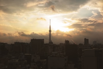 A cityscape at sunset with a tall tower, likely the Tokyo Skytree, silhouetted against a dramatic sky filled with clouds. The skyline is composed of various high-rise buildings fading into the dark as the bright sun illuminates the scene from behind the clouds.