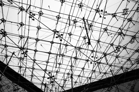 A complex structure of intersecting metal rods and wire cables form a geometric pattern against a light background. The framework appears intricate and industrial, creating an abstract and web-like appearance.