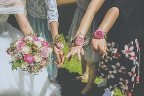 Multiple people showing their hands adorned with floral wrist corsages, accompanied by an elegant bouquet of pink and white flowers. The scene appears to be outdoors on a sunny day, indicated by the vibrant lighting and greenery.