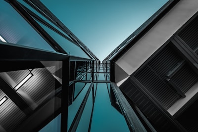A dramatic angle shot of modern architecture with glass and steel elements stretching upwards towards the sky. The geometric lines and reflections create an abstract, almost symmetrical composition, emphasizing the vertical height and sleek design.