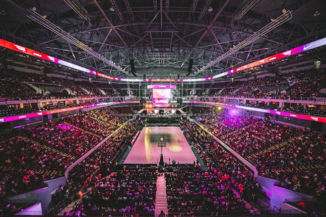 A large indoor basketball arena filled with spectators. The court is in the center, illuminated by bright lights, and surrounded by several tiers of seating. The ceiling displays an intricate web of metal beams and lighting fixtures. Electronic screens with various logos and advertisements are visible around the perimeter.