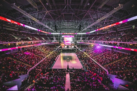 A large indoor basketball arena filled with spectators. The court is in the center, illuminated by bright lights, and surrounded by several tiers of seating. The ceiling displays an intricate web of metal beams and lighting fixtures. Electronic screens with various logos and advertisements are visible around the perimeter.