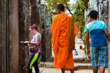 A scene at a historical site featuring two young boys and a tourist walking among ancient stone structures. One boy is wearing a traditional orange robe, often associated with religious practices, while the tourist, dressed casually, examines a book. The background includes weathered stone pillars and a glimpse of trees and vehicles in the distance.