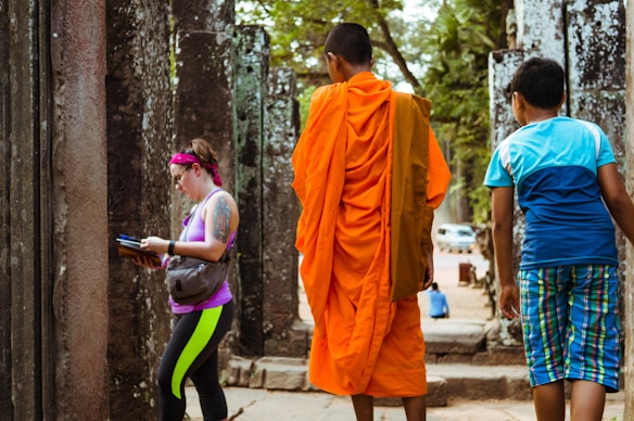 A scene at a historical site featuring two young boys and a tourist walking among ancient stone structures. One boy is wearing a traditional orange robe, often associated with religious practices, while the tourist, dressed casually, examines a book. The background includes weathered stone pillars and a glimpse of trees and vehicles in the distance.