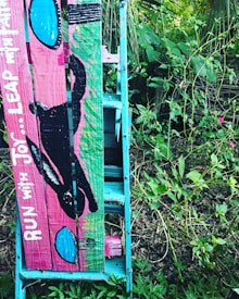 A wooden ladder-like structure painted in vibrant colors is leaning against a natural, leafy environment. The structure is painted with shades of blue, pink, and green, and features an artistic depiction of a black silhouette of a person running alongside the phrase 'RUN WITH JOY&hellip; LEAP WITH FAITH'. The surroundings are lush with green foliage and various plants.