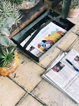 A rustic scene featuring a wooden box with colorful magazines or newspapers, a weathered tiled floor, and several potted plants. The magazines or newspapers have bright, graphic covers and one of them is placed in an open position nearby, accompanied by a blue pen.