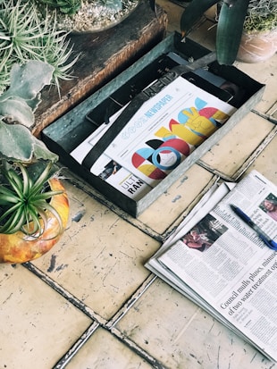 A rustic scene featuring a wooden box with colorful magazines or newspapers, a weathered tiled floor, and several potted plants. The magazines or newspapers have bright, graphic covers and one of them is placed in an open position nearby, accompanied by a blue pen.