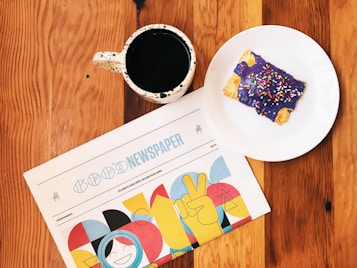 A rustic wooden table is adorned with a speckled ceramic mug filled with black coffee, set beside a plate holding a pastry covered in purple icing and colorful sprinkles. Beside them lies a newspaper titled 'Good Newspaper', featuring vibrant graphic designs with cheerful colors.
