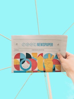 A hand is holding a newspaper with the headline 'Good Newspaper' in a modern design. The newspaper features colorful, abstract artwork with geometric shapes and a smiling face. The background is a solid light blue, creating a clean and minimalistic look.