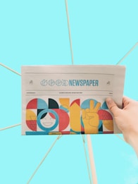 A hand is holding a newspaper with the headline 'Good Newspaper' in a modern design. The newspaper features colorful, abstract artwork with geometric shapes and a smiling face. The background is a solid light blue, creating a clean and minimalistic look.