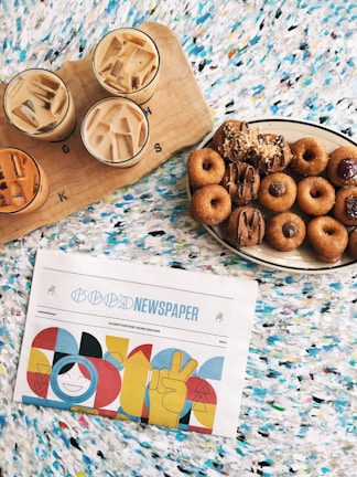 An array of iced coffee drinks served on a wooden board are paired with a plate of assorted donuts. Next to them, a newspaper with colorful, abstract designs on the cover is laid out on a vibrant, patterned surface.