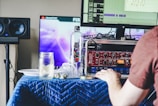 A person is working at a desk with a computer displaying an audio editing software. There are two monitors, one showing the software interface and another with a graphic image, possibly a video. The desk is covered with a blue quilted cloth, and various items including a water bottle, a jar with a drink, a plate, audio equipment with dials, and cables are strewn across it. In the background, there are speakers.