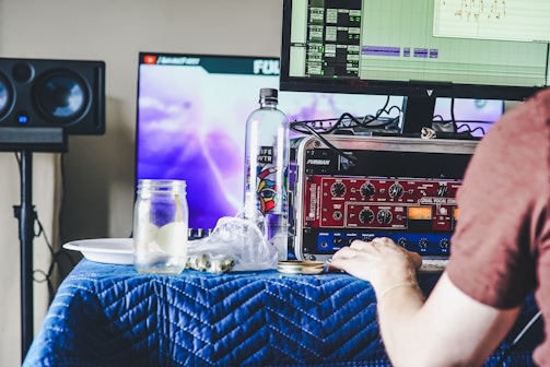 A person is working at a desk with a computer displaying an audio editing software. There are two monitors, one showing the software interface and another with a graphic image, possibly a video. The desk is covered with a blue quilted cloth, and various items including a water bottle, a jar with a drink, a plate, audio equipment with dials, and cables are strewn across it. In the background, there are speakers.