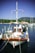 A small, colorful fishing boat is docked at a marina, flying a Greek flag. The boat is equipped with various fishing gear, ropes, and barrels on its deck. Life rings are attached to the sides, and there are several other boats in the background against a backdrop of green trees and a mountain.