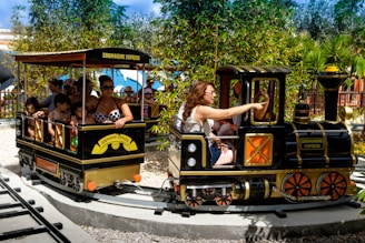 A small amusement park train is on a circular track with several passengers, including adults and children. The train is labeled 'Zoomarine Express' and features a black and yellow color scheme. It is surrounded by greenery, including bamboo and other plants. The adults and children appear to be enjoying the ride on a sunny day.