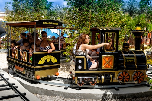 A small amusement park train is on a circular track with several passengers, including adults and children. The train is labeled 'Zoomarine Express' and features a black and yellow color scheme. It is surrounded by greenery, including bamboo and other plants. The adults and children appear to be enjoying the ride on a sunny day.