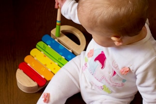 A baby is sitting on a wooden floor, wearing a white outfit with colorful patterns. In front of the baby is a rainbow-colored toy xylophone. The baby holds a mallet and appears to be playing with the xylophone.