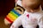 A baby is sitting on a wooden floor, wearing a white outfit with colorful patterns. In front of the baby is a rainbow-colored toy xylophone. The baby holds a mallet and appears to be playing with the xylophone.